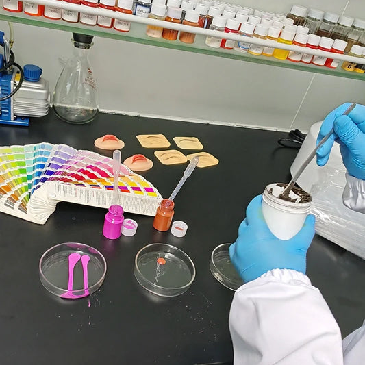 Tattoo pigment color testing in a laboratory with color charts, sample pigments, and gloved hands mixing ink.
