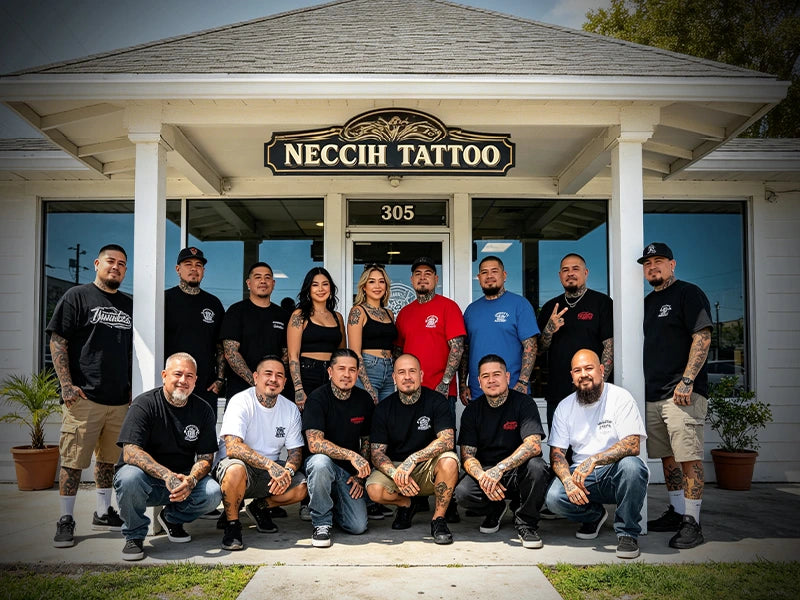 A group of 15 tattoo artists and staff members posing in front of their storefront, 'Neccih Tattoo', wearing matching black and white t-shirts with the brand logo.