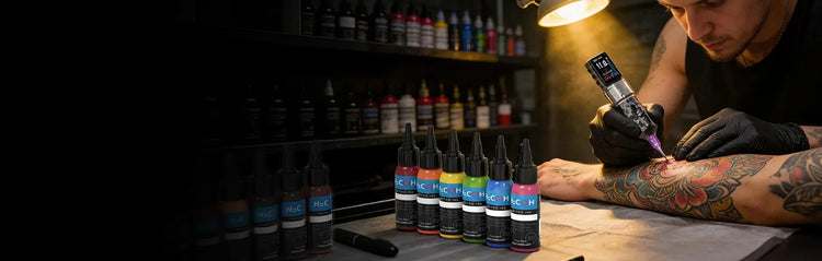A tattoo artist wearing black gloves applies a tattoo to a client's forearm using a tattoo machine under warm overhead lighting. Several neccih ink bottles with colorful labels are lined up on the table in the foreground, with shelves of more bottles blurred in the dark background.