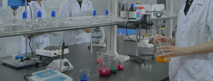 A laboratory scene with multiple people in white lab coats working at a bench with glassware, test tubes, a microscope, and other scientific equipment. The image is heavily darkened and low-contrast.