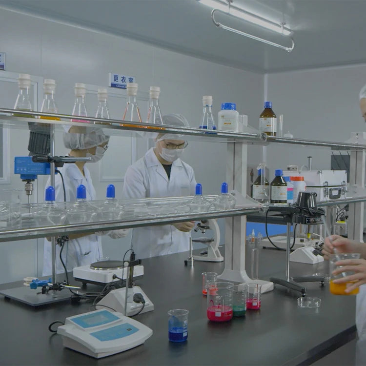 A dimly lit laboratory scene with a person in a white lab coat, face mask, and hair cover standing behind a workbench with glassware, test tubes, beakers, and scientific equipment.